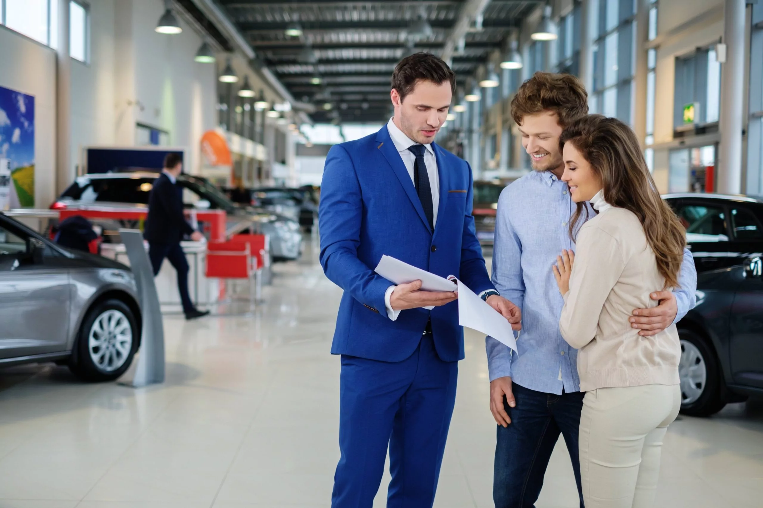 Car dealership representative discussing financing options with a couple in a modern showroom, surrounded by quality used cars, emphasizing a friendly and transparent buying experience.