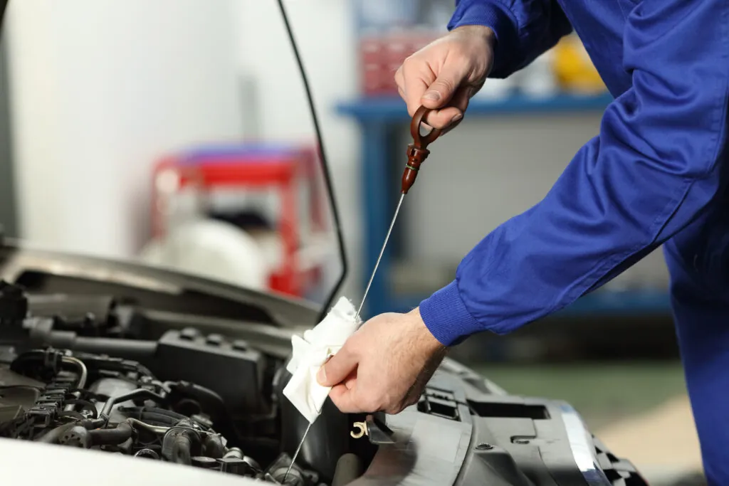 Person in blue coveralls checking car oil level with dipstick and cloth under the hood of a vehicle, illustrating car maintenance importance.
